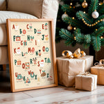 A framed Christmas alphabet art print leaning against a beige sofa on a wooden floor, next to wrapped gift boxes and a decorated Christmas tree.