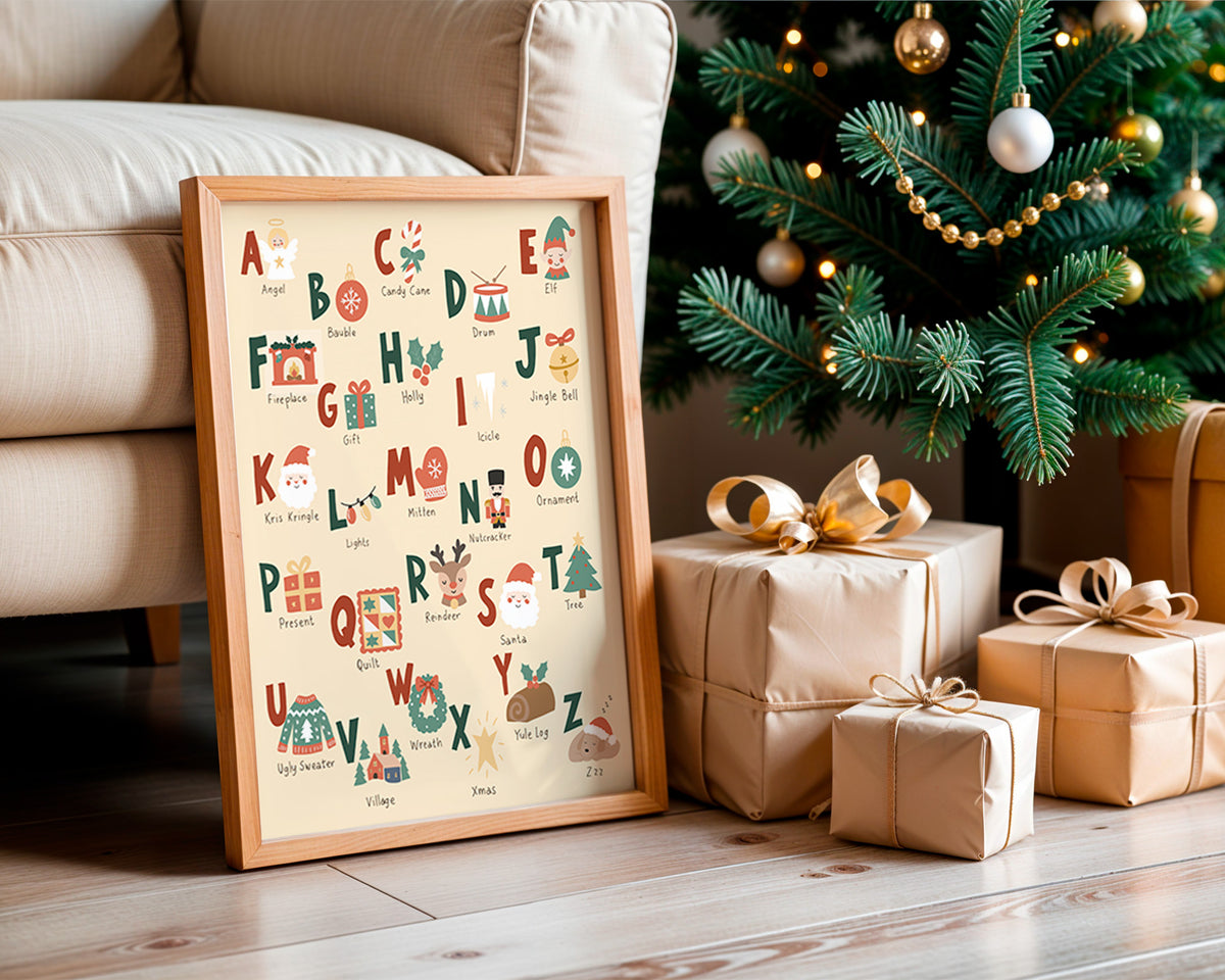 A framed Christmas alphabet art print leaning against a beige sofa on a wooden floor, next to wrapped gift boxes and a decorated Christmas tree.