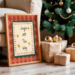 A framed Christmas art print leaning against a beige sofa on a wooden floor, next to wrapped gift boxes and a decorated Christmas tree. The print features the text 'Jingle All The Way' with gold stars on a cream background, bordered by a green scalloped frame and an orange argyle patterned border.