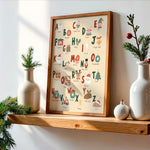 A framed alphabet art print featuring Christmas-themed illustrations and names, displayed on a wooden shelf in a styled home setting with white ceramic vases and pine branches.