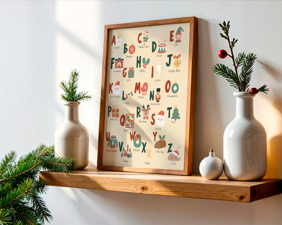 A framed alphabet art print featuring Christmas-themed illustrations and names, displayed on a wooden shelf in a styled home setting with white ceramic vases and pine branches.