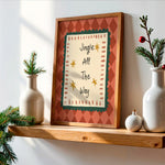 Framed wall art with the phrase 'Jingle All The Way' in a rustic wood frame, styled on a wooden shelf with white ceramic vases, evergreen branches, and small ornaments against a white wall.
