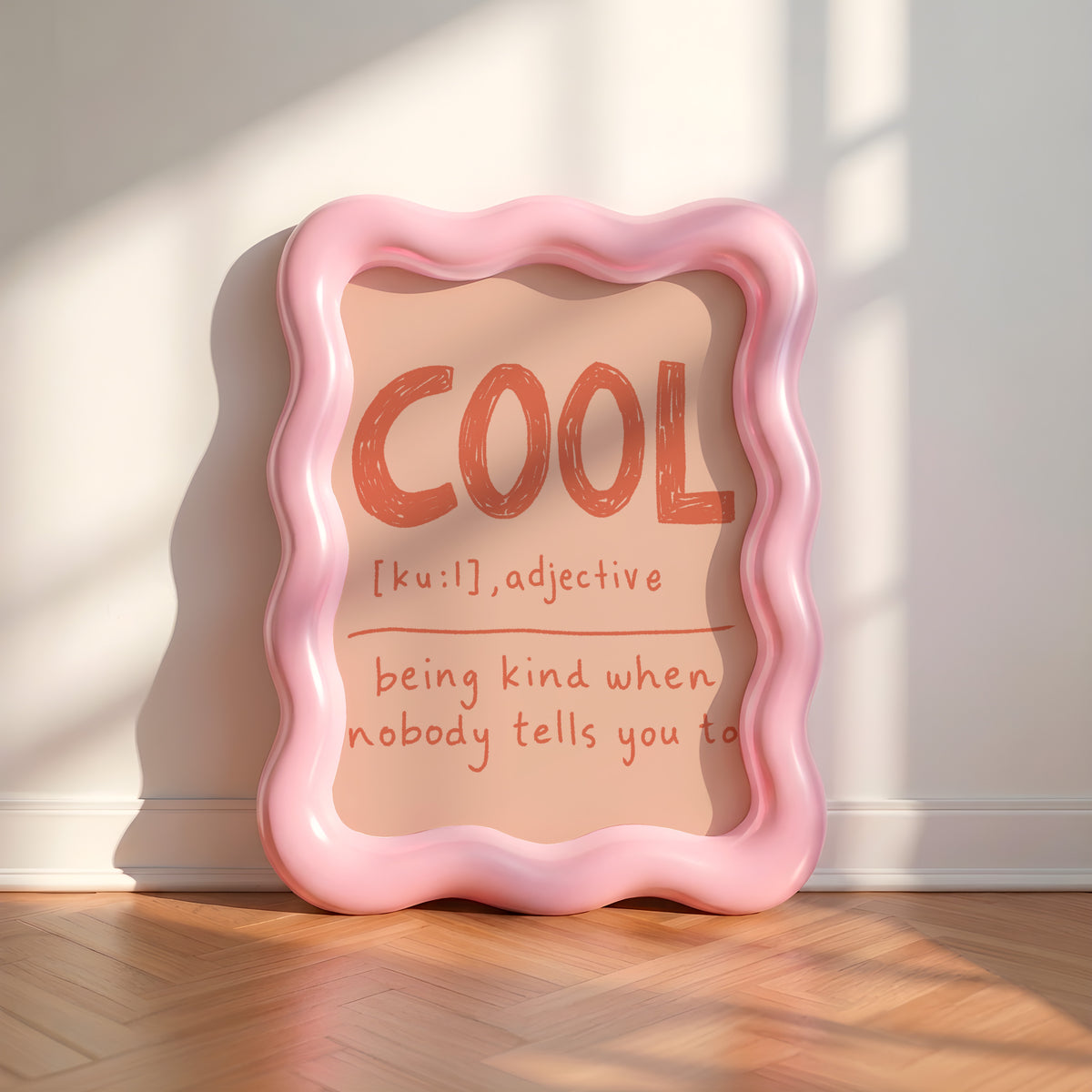 A wavy pink decorative frame leaning against a white wall on a wood floor, with the word "COOL" and a dictionary-style definition printed inside in warm orange-red text.