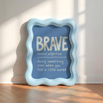 A decorative framed wall sign with a wavy light blue border and a blue center panel, leaning against a white wall on a wood floor. The panel reads “BRAVE” with a dictionary-style definition underneath in white text.