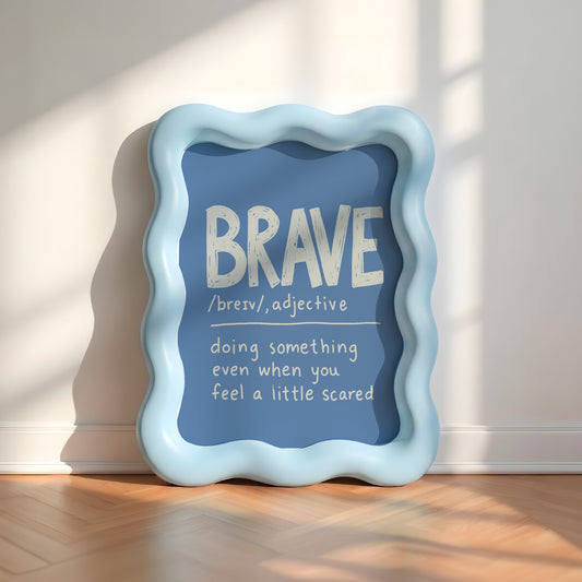 A decorative framed wall sign with a wavy light blue border and a blue center panel, leaning against a white wall on a wood floor. The panel reads “BRAVE” with a dictionary-style definition underneath in white text.
