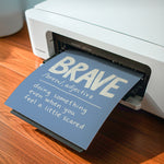 A printed card or poster with the word “BRAVE” and a definition slides out of a white printer on a wooden desk.