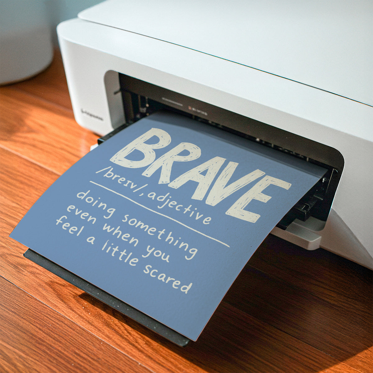 A printed card or poster with the word “BRAVE” and a definition slides out of a white printer on a wooden desk.