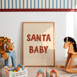 A framed art print with the text 'SANTA BABY' in bold red letters on a cream background, leaning against a white wall in a styled children's room with toys and a rocking horse.