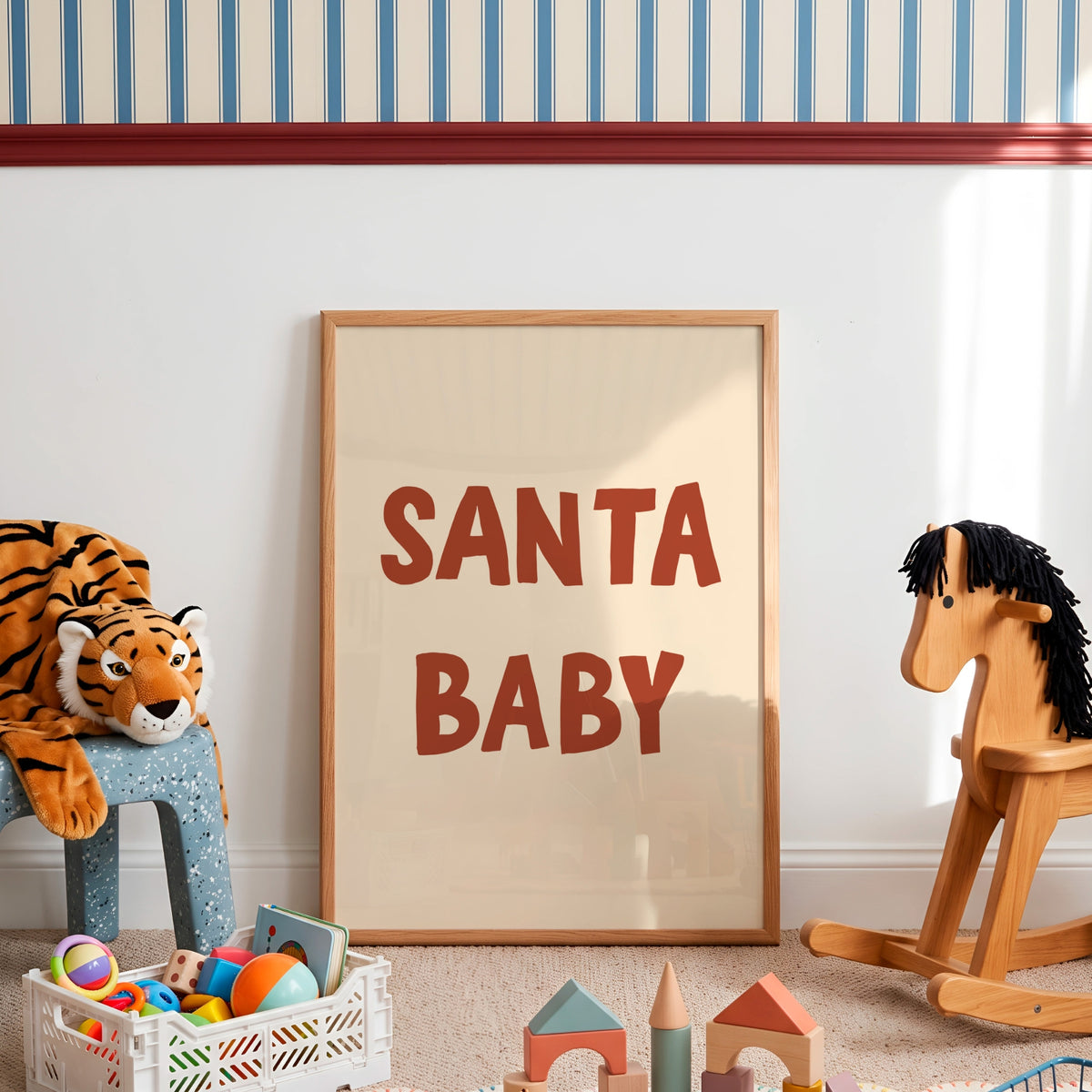 A framed art print with the text 'SANTA BABY' in bold red letters on a cream background, leaning against a white wall in a styled children's room with toys and a rocking horse.
