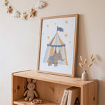 A framed nursery art print featuring a cartoon elephant under a colorful circus tent, styled on a wooden shelf in a warm, neutral-toned room with a plush bunny, books, and a small vase.
