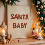 A framed Christmas sign reading 'SANTA BABY' in dark red letters on a cream background, styled on a wooden shelf with holiday greenery, silver ornaments, lanterns, and a nutcracker.