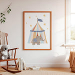 A framed nursery art print featuring a cartoon elephant under a striped circus tent, hung on a white wall in a softly styled child's room with a rocking chair, books, and a window.