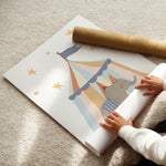 A person is unrolling a large white wall mural featuring a whimsical circus tent with a gray elephant inside, gold stars, and a brown cardboard roll on top, all laid out on a beige carpet.
