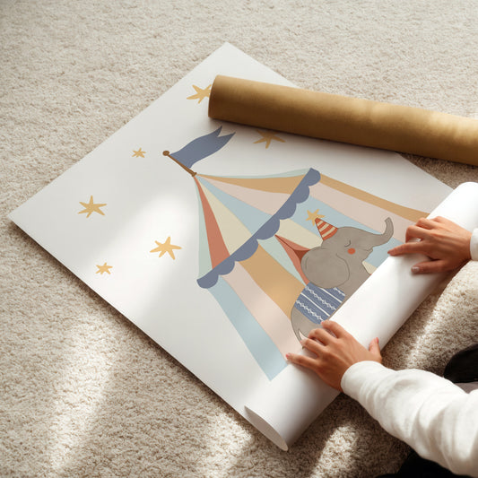 A person is unrolling a large white wall mural featuring a whimsical circus tent with a gray elephant inside, gold stars, and a brown cardboard roll on top, all laid out on a beige carpet.