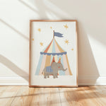 A framed children's art print leaning against a white wall on a light wood floor. The print shows a gray elephant wearing a party hat under a pastel striped circus tent with gold stars.
