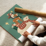 A person unrolls a large sheet of Christmas-themed wrapping paper on a beige carpet, with a rolled gold tube of paper beside it. The sheet features a festive illustration of a polar bear, deer, snowman, and stars in red, brown, white, and teal.