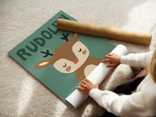 A person unrolls a large printed poster or wall decal featuring a cartoon reindeer and the name 'RUDOLPH' on a teal background, placed on a beige carpet. A rolled brown paper sheet lies nearby.
