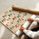 A person unrolls a Christmas-themed alphabet wrapping paper on a beige carpet, with a gold cardboard mailing tube placed nearby.