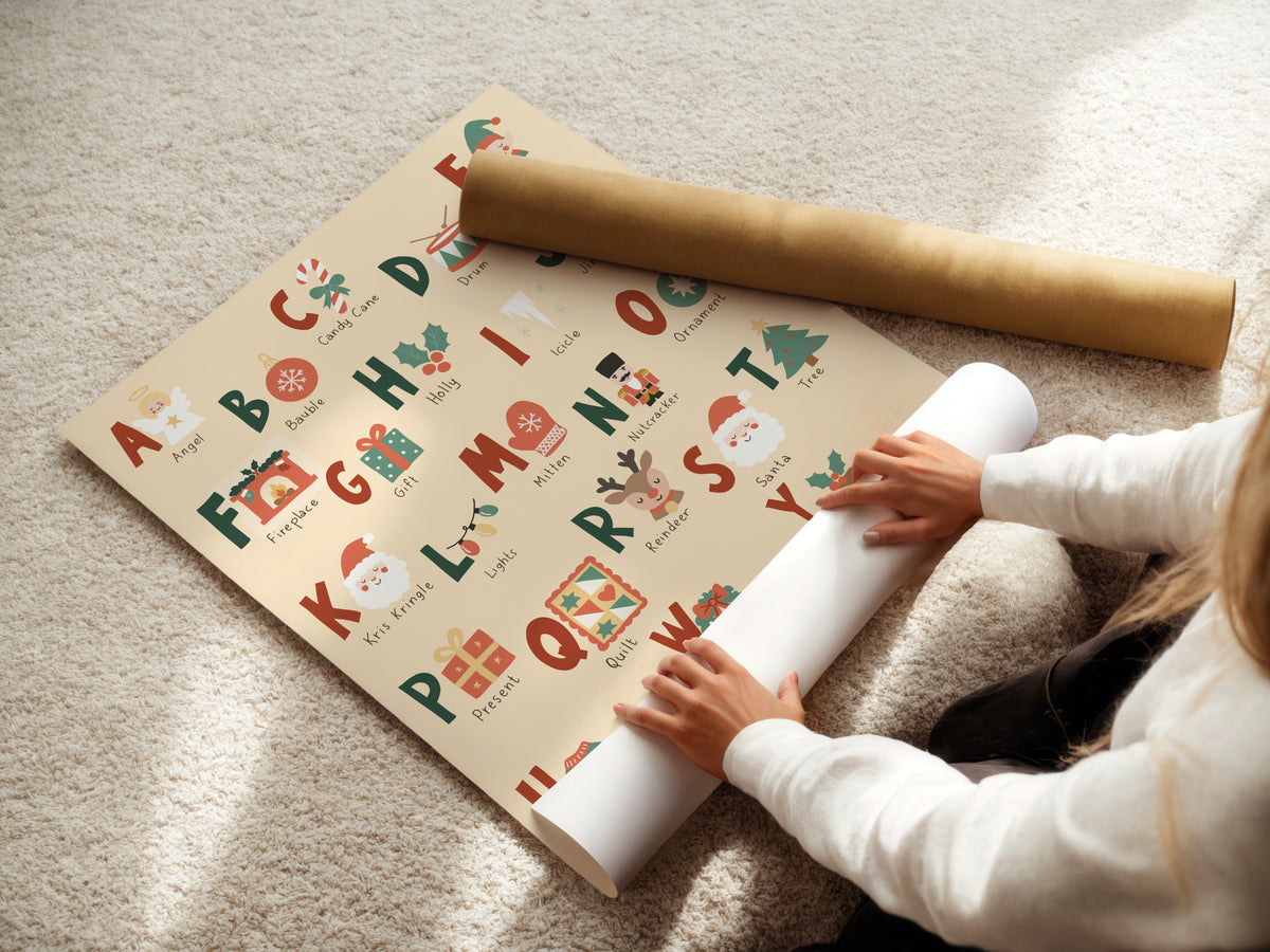 A person unrolls a Christmas-themed alphabet wrapping paper on a beige carpet, with a gold cardboard mailing tube placed nearby.