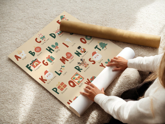 A person unrolls a Christmas-themed alphabet wrapping paper on a beige carpet, with a gold cardboard mailing tube placed nearby.
