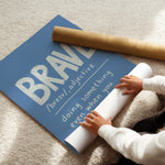 A person unrolling a large blue wall art print on a beige carpet, with a brown cardboard mailing tube nearby. The print features the word 'BRAVE' in large white letters with pronunciation guidance below it.