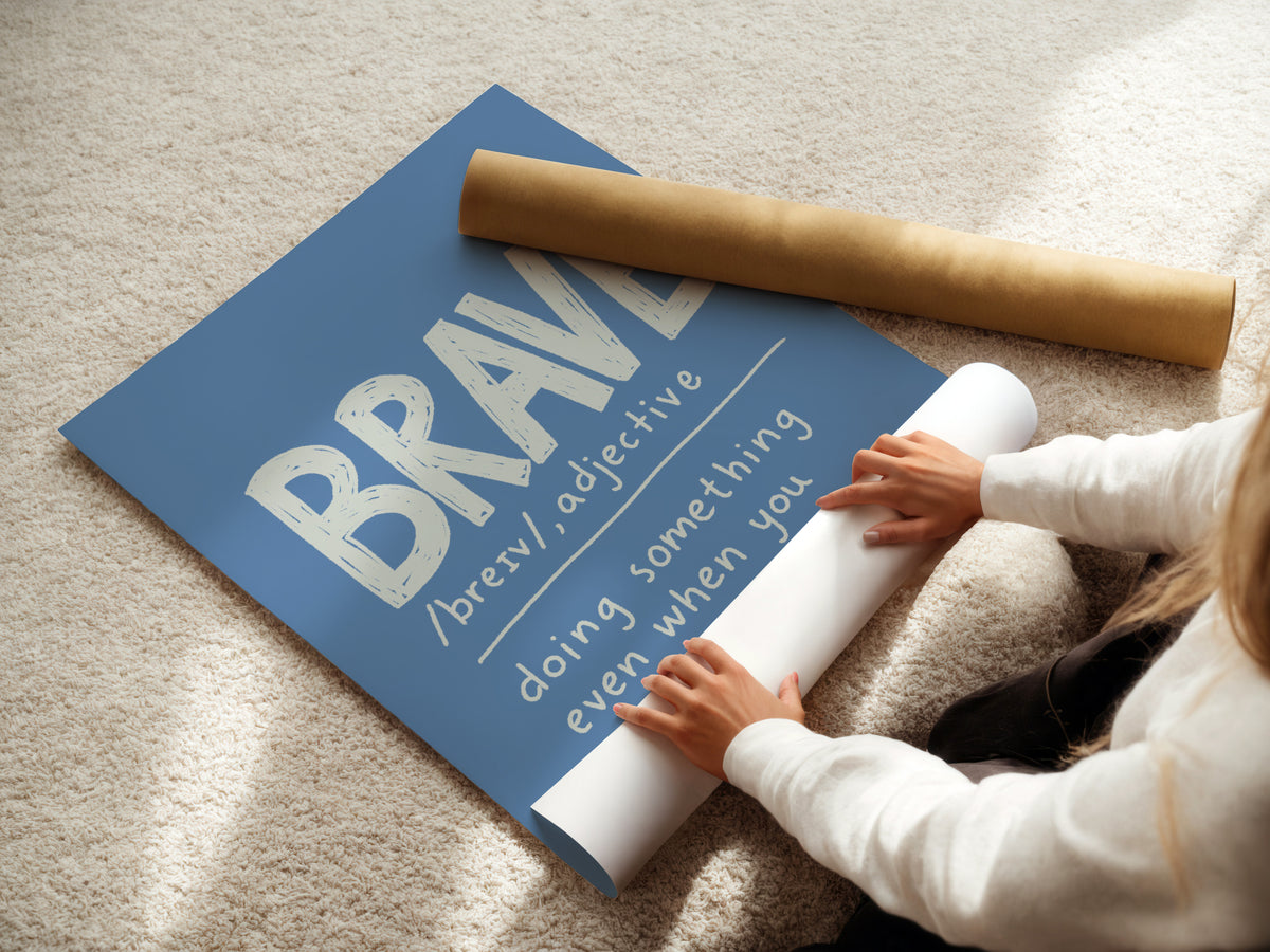 A person unrolling a large blue wall art print on a beige carpet, with a brown cardboard mailing tube nearby. The print features the word 'BRAVE' in large white letters with pronunciation guidance below it.