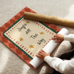 A person unrolls a decorative holiday poster on a beige carpet, with the printed message 'Jingle All The Way' visible and a rolled gold paper tube beside it.