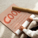 A person unrolls a large peach-colored poster or mat on a beige carpet, with a rolled kraft paper tube placed across the top. The printed design features the word "COOL" in large orange-red letters and handwritten-style text underneath.