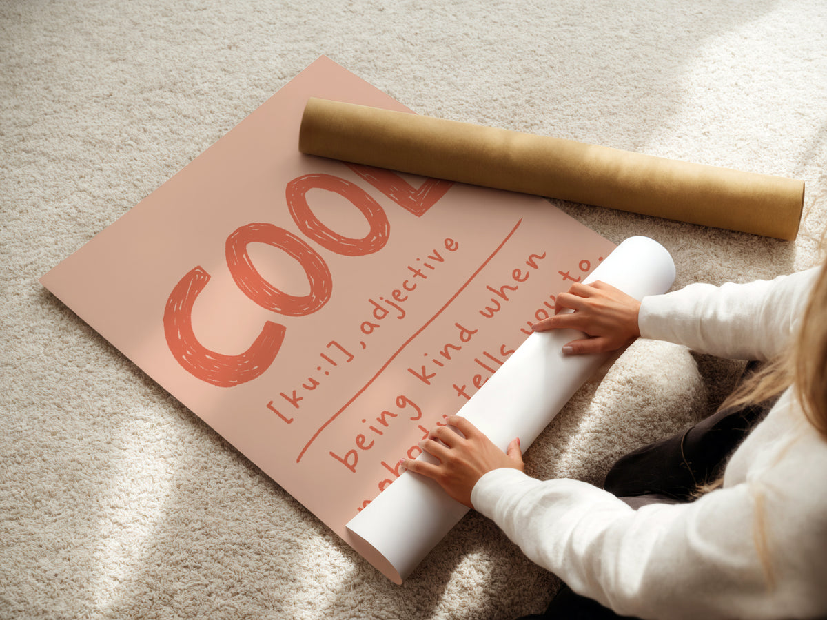 A person unrolls a large peach-colored poster or mat on a beige carpet, with a rolled kraft paper tube placed across the top. The printed design features the word "COOL" in large orange-red letters and handwritten-style text underneath.