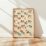 A framed alphabet art print featuring Christmas-themed illustrations and words, leaning against a white wall on a light wood floor with soft diagonal shadows.