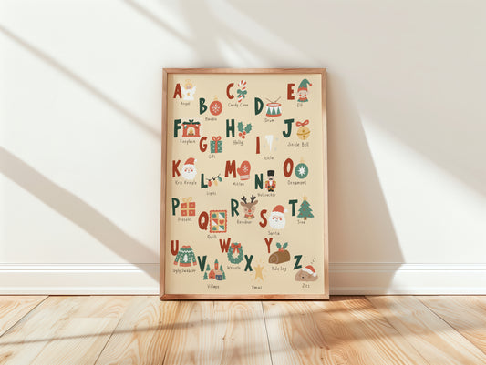 A framed alphabet art print featuring Christmas-themed illustrations and words, leaning against a white wall on a light wood floor with soft diagonal shadows.