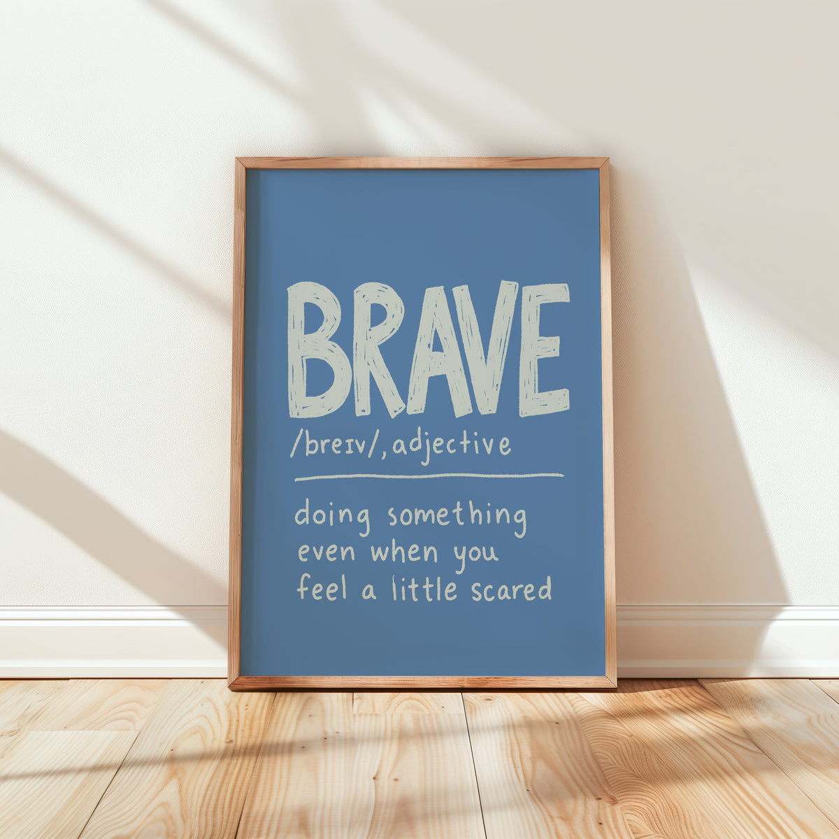 A framed typographic wall art print leaning against a white wall on a light wood floor. The print has a muted blue background with white text reading “BRAVE /breiv/, adjective” and a definition below, surrounded by a natural wood frame.