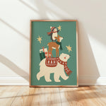 A framed art print leaning against a white wall on a light wood floor. The print shows a whimsical illustration of a polar bear wearing a red patterned scarf and carrying a deer on its back, with a penguin, stars, and gift bags on a muted teal background.