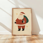 A framed art print featuring a cute, minimalist cartoon illustration of Santa Claus standing on a light wood floor against a white wall with soft window shadows.