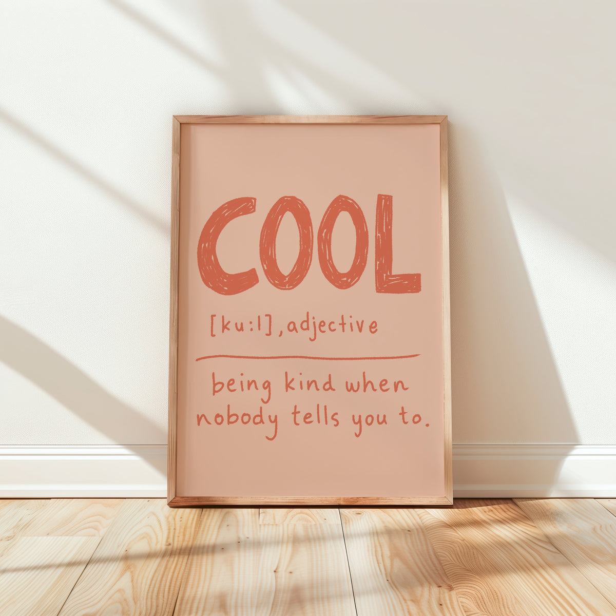 A framed typographic wall art print leaning against a white wall on a light wood floor. The print has a peach background with the word "COOL" in large textured orange-red letters and smaller handwritten-style text underneath reading "[ku:ɪl], adjective being kind when nobody tells you to.