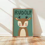 A framed children's art print featuring a cute cartoon reindeer with the name 'RUDOLF' above it, leaning against a white wall on a light wood floor with soft window shadows.