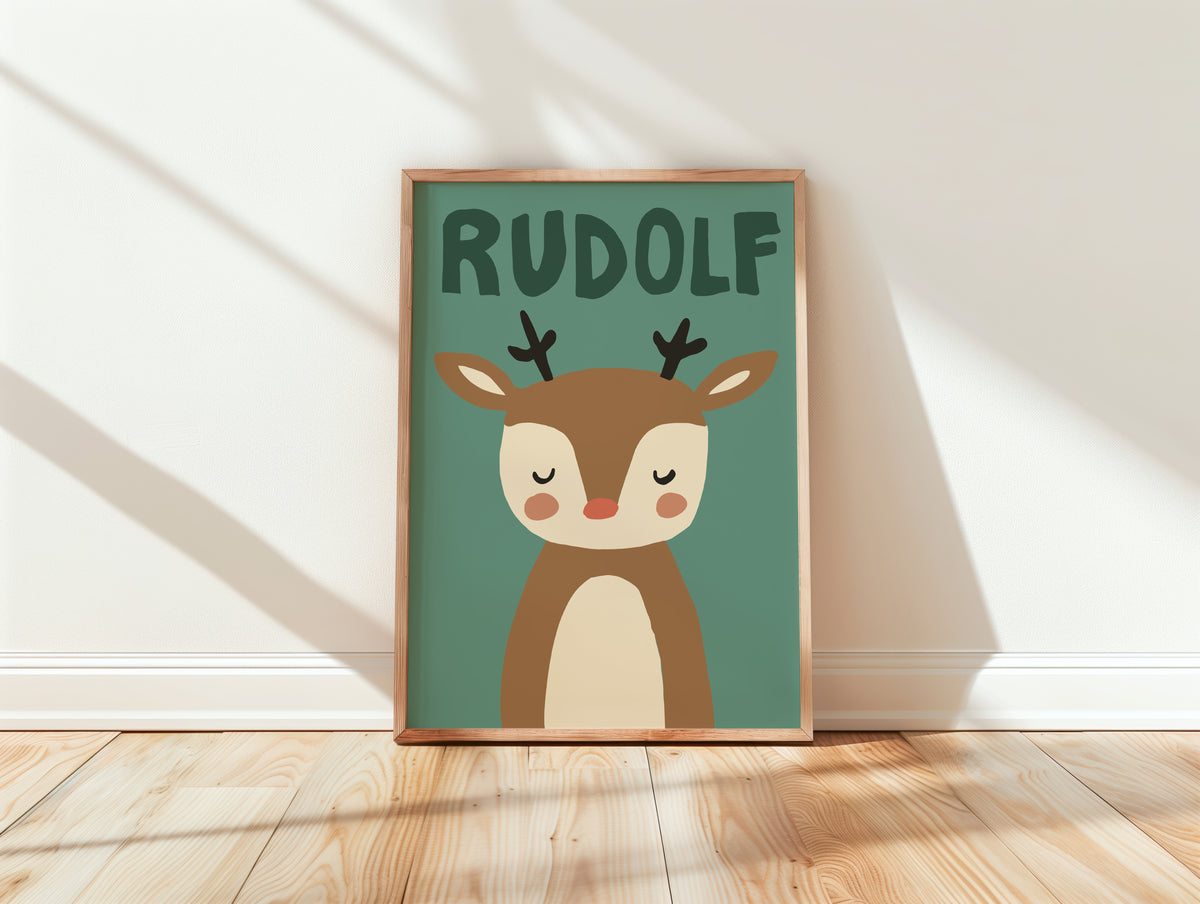 A framed children's art print featuring a cute cartoon reindeer with the name 'RUDOLF' above it, leaning against a white wall on a light wood floor with soft window shadows.