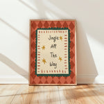 A framed holiday art print leaning against a white wall on a light wood floor. The print features the text 'Jingle All The Way' in a playful handwritten font, surrounded by a cream center panel with a dark green scalloped border, yellow stars, and a terracotta orange diamond-patterned frame.